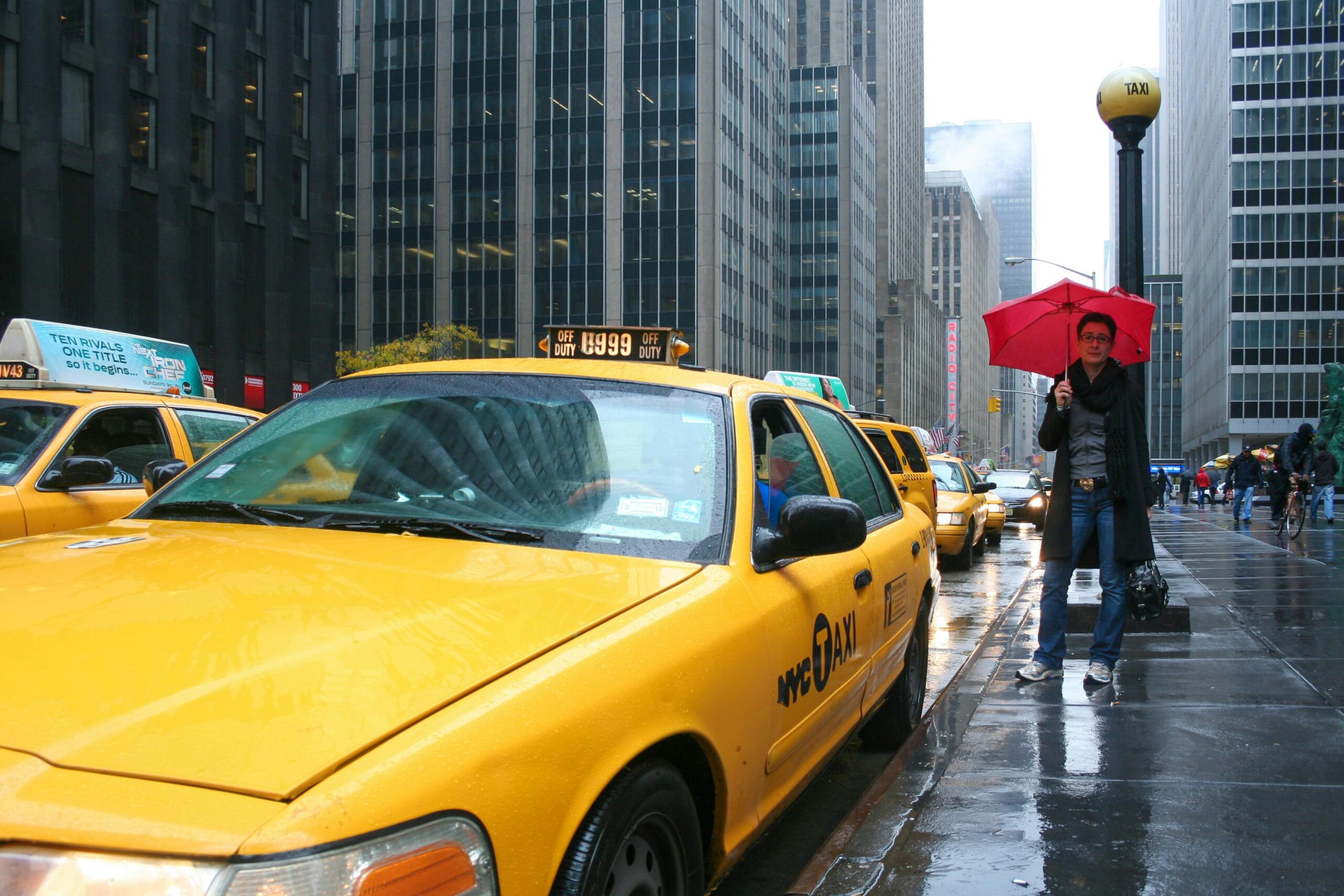 Umbrella-wielding pedestrian on wet New York street surrounded by yellow taxis and skyscrapers.