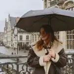 A woman in warm clothing holds an umbrella on a rainy day in a European city, with historic architecture and a river view.
