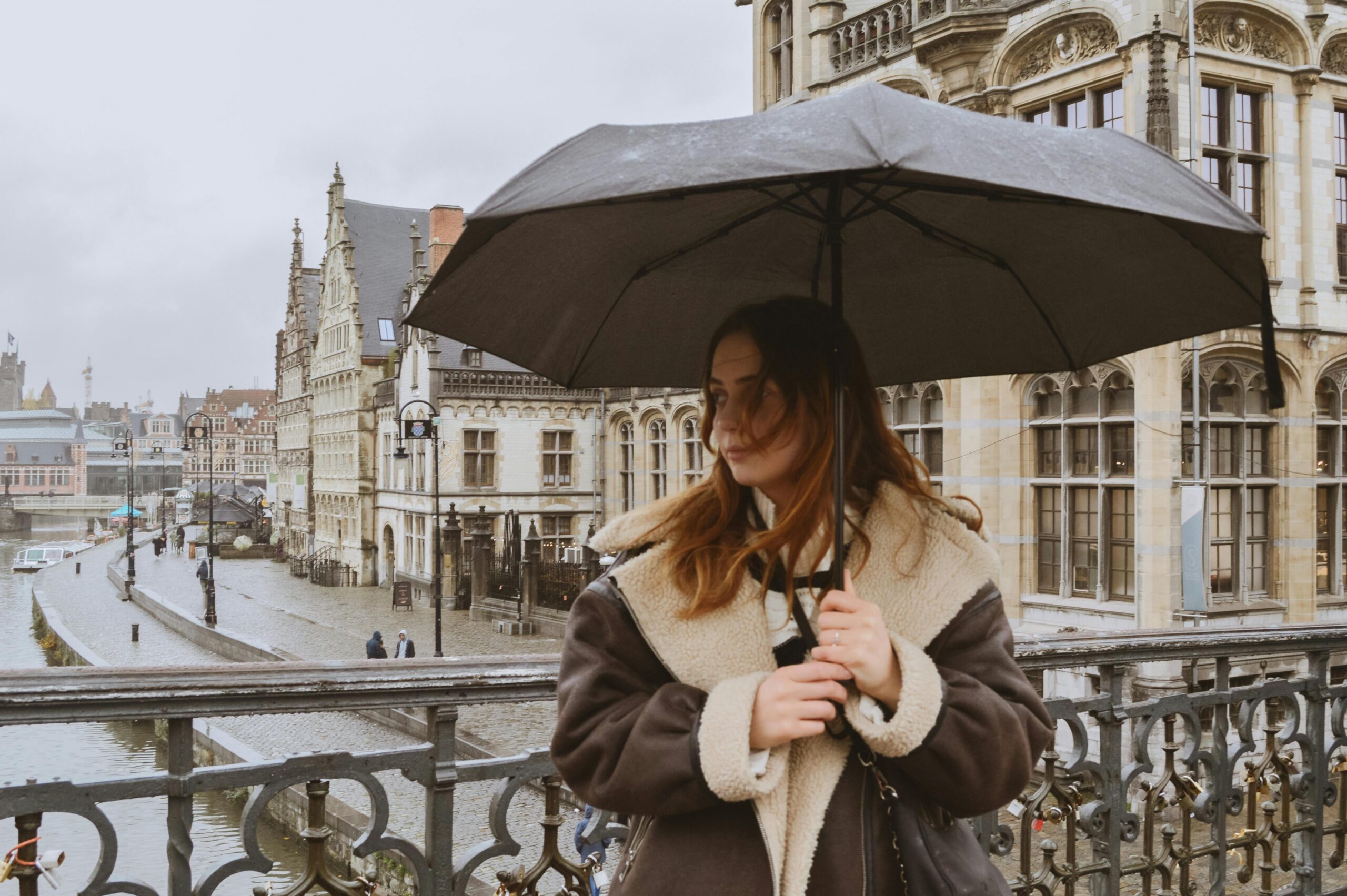 A woman in warm clothing holds an umbrella on a rainy day in a European city, with historic architecture and a river view.